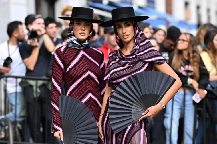 Two guests in pleated stripes, wide-brimmed hats and hand fans photographed outside a show at Milan Fashion Week, an engagement in the season calendar of The Mansa Court