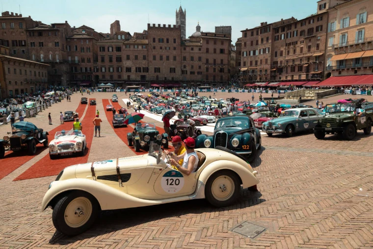 A cream pre-war roadster crosses the brick of the Piazza del Campo in Siena, the field of classic cars gathered behind. The Mille Miglia, from Brescia to Rome and back, in the season calendar of The Mansa Court