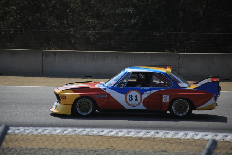 A BMW coupe racing car painted in bold red, blue and yellow art-car livery, number 31, speeds past the track wall at Laguna Seca (2017).