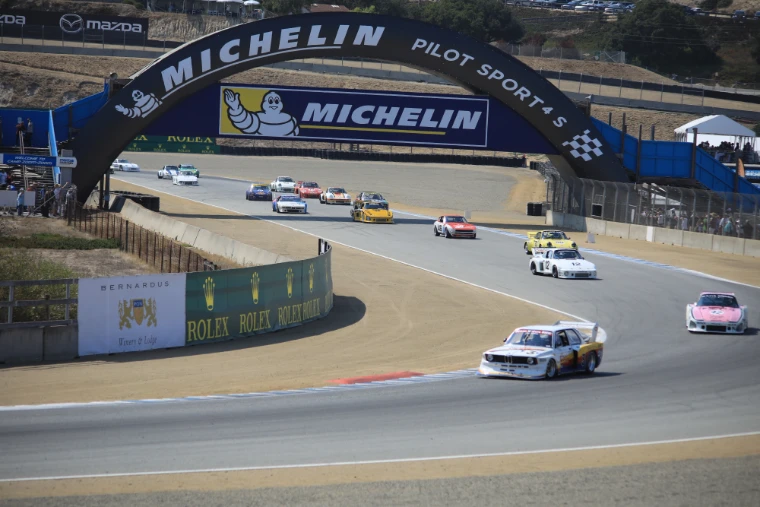 A long line of historic racing cars streams downhill beneath the arched Michelin bridge at Laguna Seca, with Rolex banners along the track wall (2017).