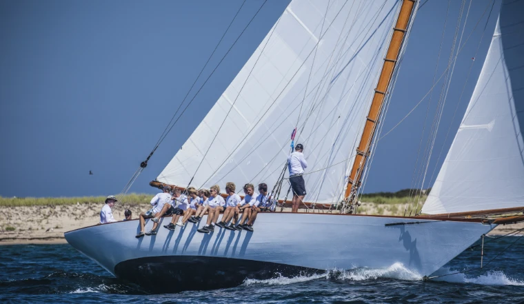 A crew in white shirts sits along the windward rail of a heeling classic yacht, its pale blue hull cutting through dark blue water off a sandy shore (2015).
