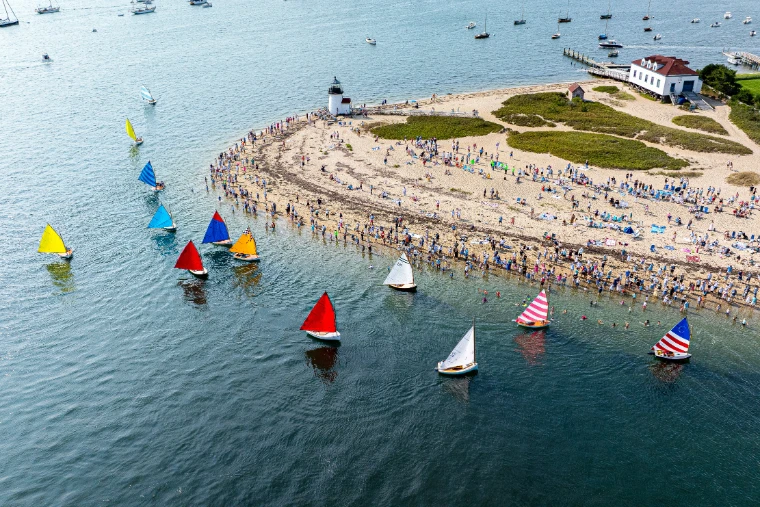 Aerial view of small catboats with brightly coloured sails rounding Brant Point lighthouse as crowds line the beach (2025). Nantucket Race Week, Brant Point, Nantucket Harbor, Massachusetts, in the season calendar of The Mansa Court