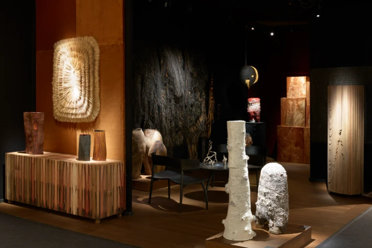 Ceramic and carved-wood objects on plinths and a low cabinet in a softly lit gallery stand with dark walls (2023).