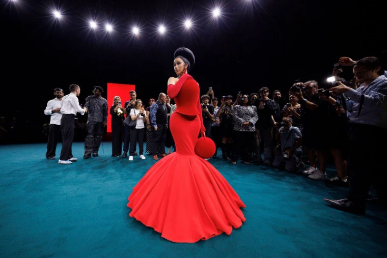 A guest in a scarlet fishtail gown turning for a bank of photographers on the teal carpet at Paris Fashion Week, an engagement in the season calendar of The Mansa Court