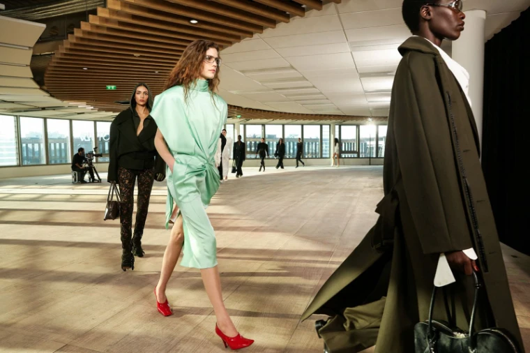 Models in tailoring and silk circling a daylit upper floor with windows onto the city at Paris Fashion Week, an engagement in the season calendar of The Mansa Court
