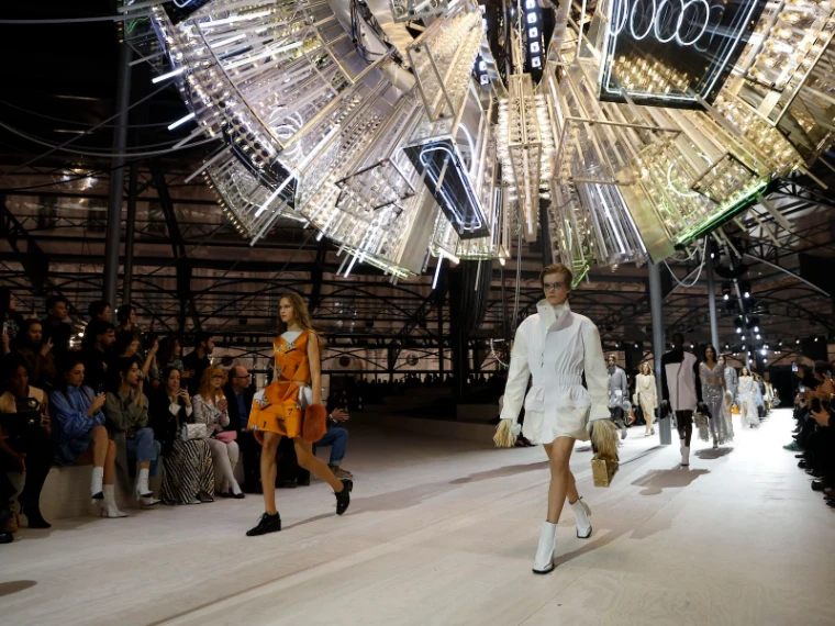 Models passing along a pale runway beneath an enormous suspended sculpture of mirrors and neon tubes at Paris Fashion Week, an engagement in the season calendar of The Mansa Court