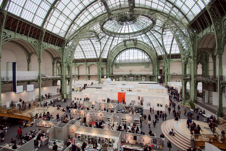 The nave of the Grand Palais seen from the gallery during Paris Photo, rows of white gallery stands and visitors beneath the green ironwork and glass roof (2016).