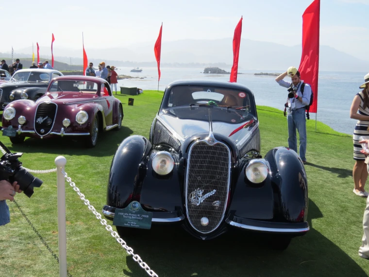 Two streamlined Alfa Romeo coupes, one black and one maroon, stand roped off on the lawn beside Carmel Bay under red pennants as photographers and spectators look on (2015).