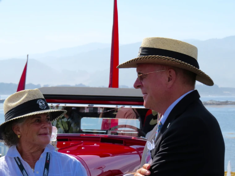 A man and a woman in straw hats talk beside a red open car on the show lawn, with the water and hazy hills behind them (2015).