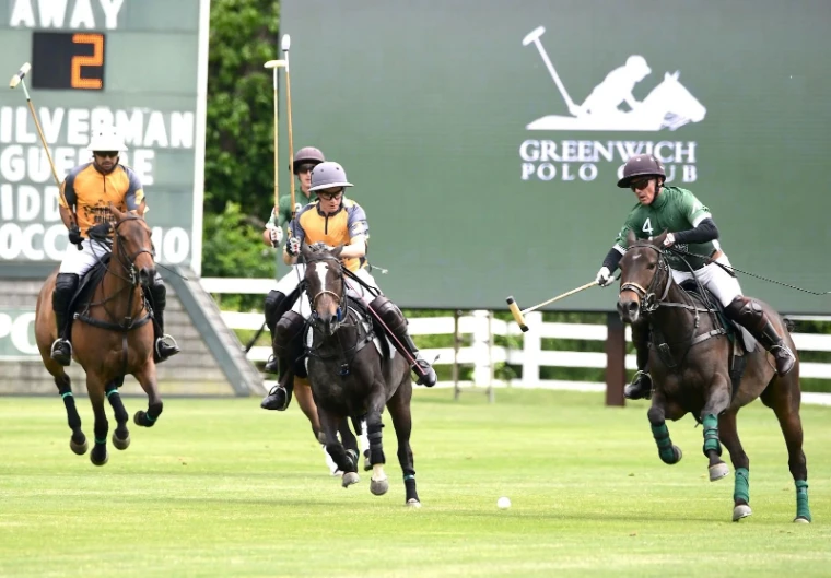 Three players in orange and green shirts galloping after the ball before the Greenwich Polo Club scoreboard, for Polo in Greenwich, Connecticut, a summer afternoon in the season calendar of The Mansa Court