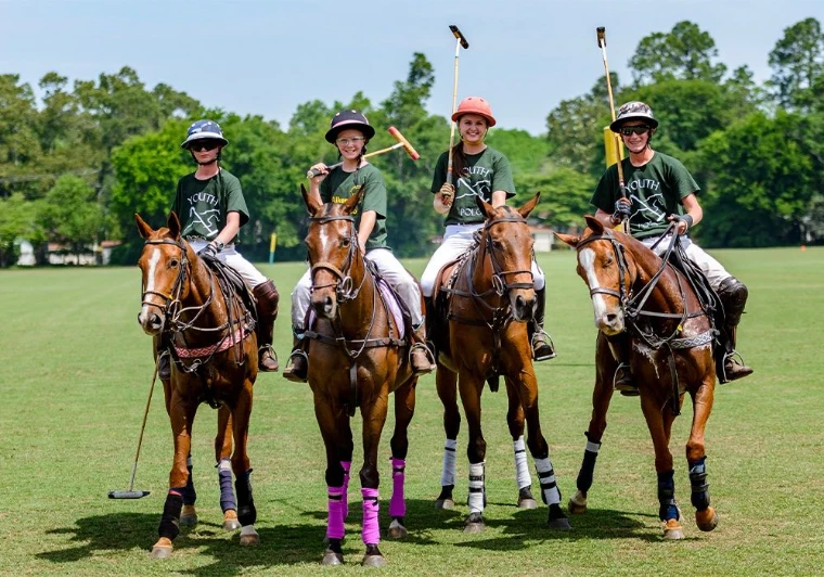 Four young riders in helmets and green shirts lined up on polo ponies with their mallets raised, on a grass field before trees, for Polo Lessons and Equestrian Riding, Bridgehampton, New York, a morning in the season calendar of The Mansa Court