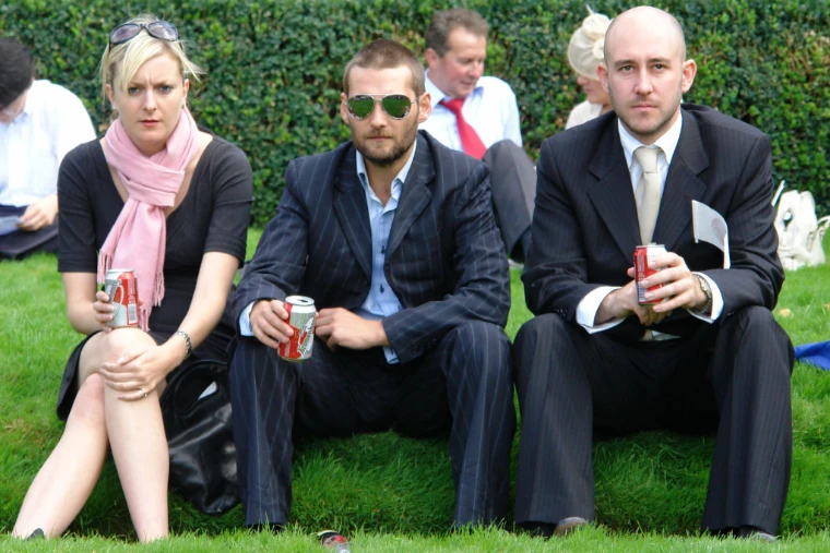 Three racegoers, two men in dark suits and a woman with a pink scarf, sitting on the grass before a hedge with drinks in hand (2006).