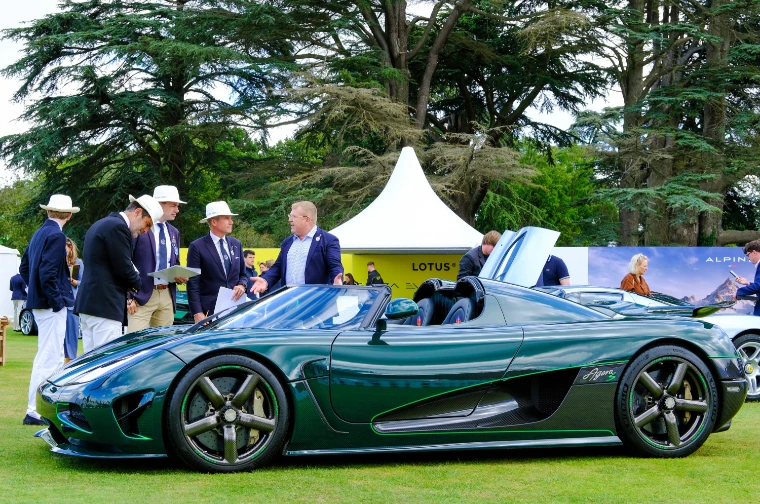 Men in blazers and panama hats, several holding clipboards, gather around a dark green open-topped Koenigsegg on the lawn beside white marquees (2023).