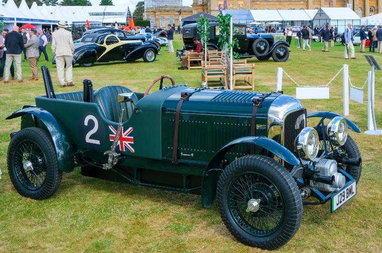 A green vintage Bentley racing car with the number 2 and a Union flag on its flank stands on the grass, with other pre-war cars and visitors behind (2023).