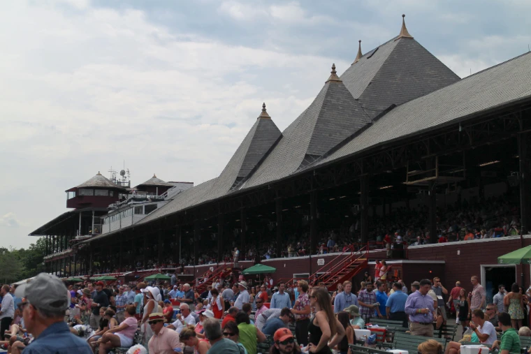 Racegoers fill the lawn and benches beneath the slate-roofed, spired grandstand on a summer race day (2015). Saratoga Race Course, Saratoga Springs, New York, shown for Saratoga Opening Weekend in the season calendar of The Mansa Court