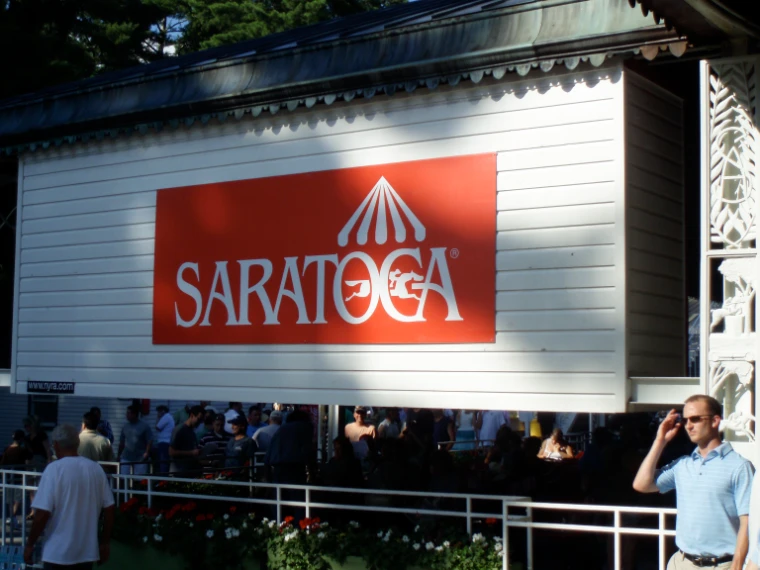 Racegoers gather in the shade beneath a white clapboard stand bearing a red Saratoga sign (2008). Saratoga Race Course, Saratoga Springs, New York, shown for Saratoga Opening Weekend in the season calendar of The Mansa Court