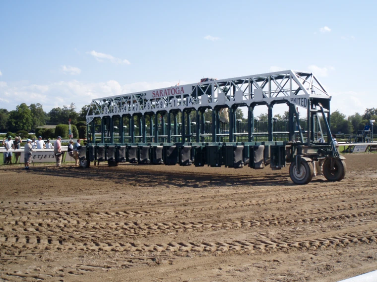 The green Saratoga starting gate stands on the raked dirt track under a clear blue sky (2008). Saratoga Race Course, Saratoga Springs, New York, shown for Saratoga Opening Weekend in the season calendar of The Mansa Court