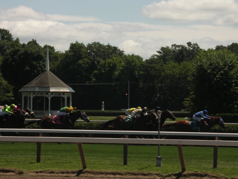 Horses race on the turf past the white gazebo of the infield, framed by tall summer trees (2005). Saratoga Race Course, Saratoga Springs, New York, shown for Saratoga Opening Weekend in the season calendar of The Mansa Court