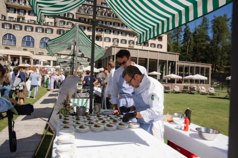 A guest chef finishing rows of tartare plates under a green-striped awning on the hotel lawn at the St. Moritz Gourmet Festival, an engagement in the season calendar of The Mansa Court