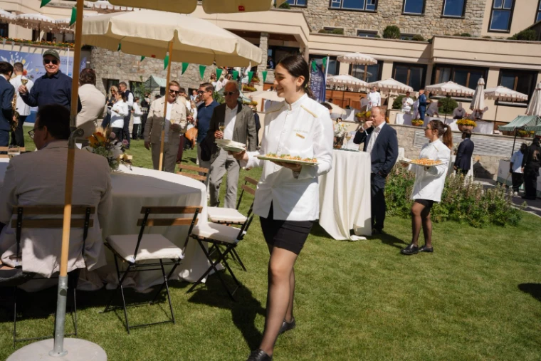 Staff in white jackets carrying trays of canapés across the hotel lawn at the St. Moritz Gourmet Festival garden party, an engagement in the season calendar of The Mansa Court