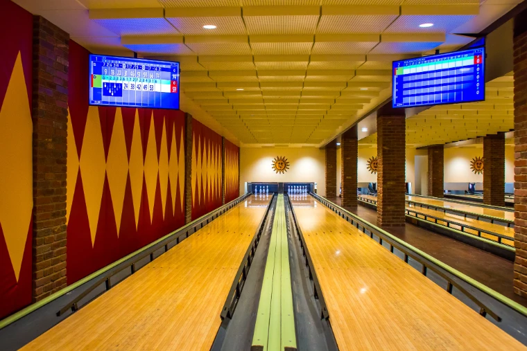 Polished wooden lanes of a small bowling alley with red-and-yellow diamond walls, brick pillars, sun-face emblems and overhead scoreboards (2015).