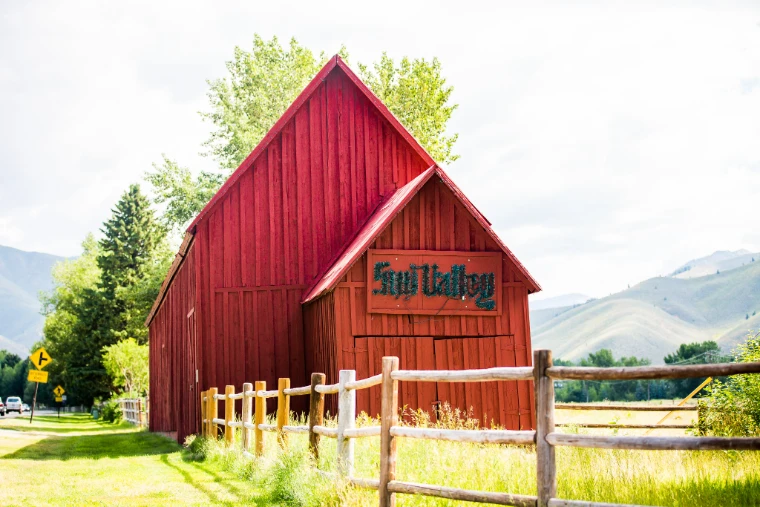 A red wooden barn with a green “Sun Valley” sign behind a split-rail fence, with summer grass, leafy trees and green hills beyond (2015).