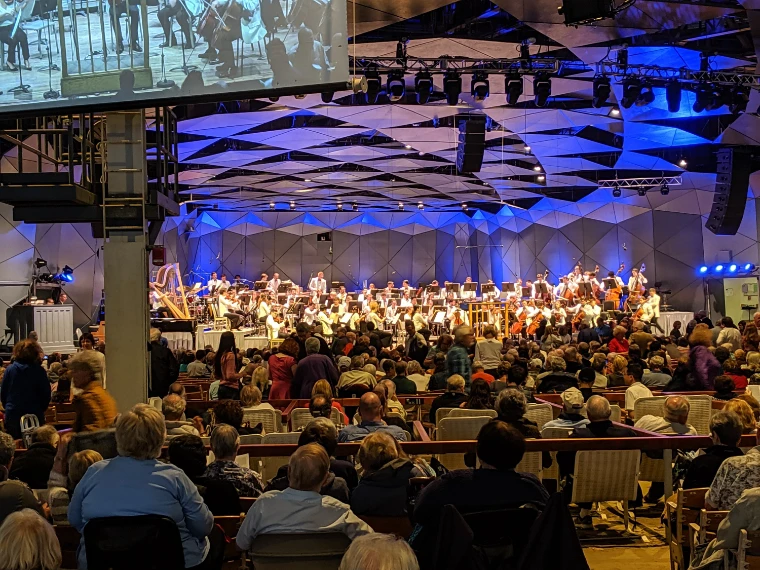 An orchestra in white jackets performs on the blue-lit stage of the Music Shed beneath its angular acoustic canopy, watched by a packed audience and a large video screen (2019).