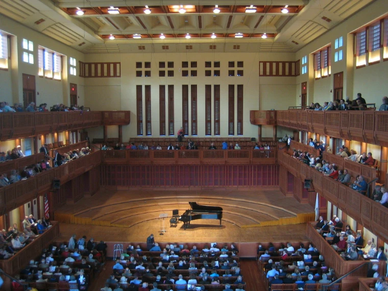 A warm wood-panelled concert hall with tiered balconies and tall slatted windows looks down on a grand piano lit on stage as the audience takes its seats (2009).