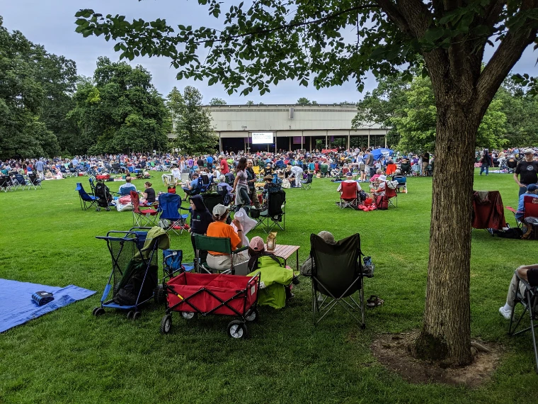 Concertgoers with folding chairs, wagons and picnic tables fill the broad lawn under shade trees, with the open-sided Music Shed beyond (2019).