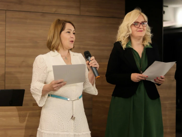 Two women reading from sheets of notes before a slatted wooden wall, one in white speaking into a hand microphone, for The Acquisition, New York, mid to late April in the season calendar of The Mansa Court