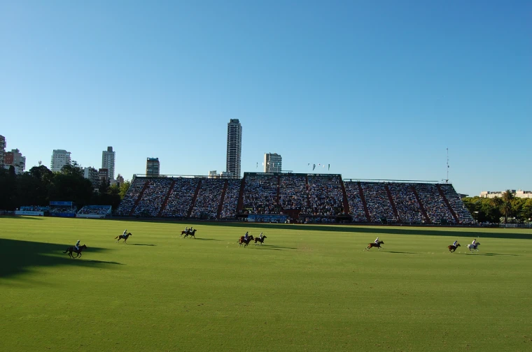 Players spread across the main ground in a match of the Argentine Open, the full main stand and city towers behind under a clear sky (2012).