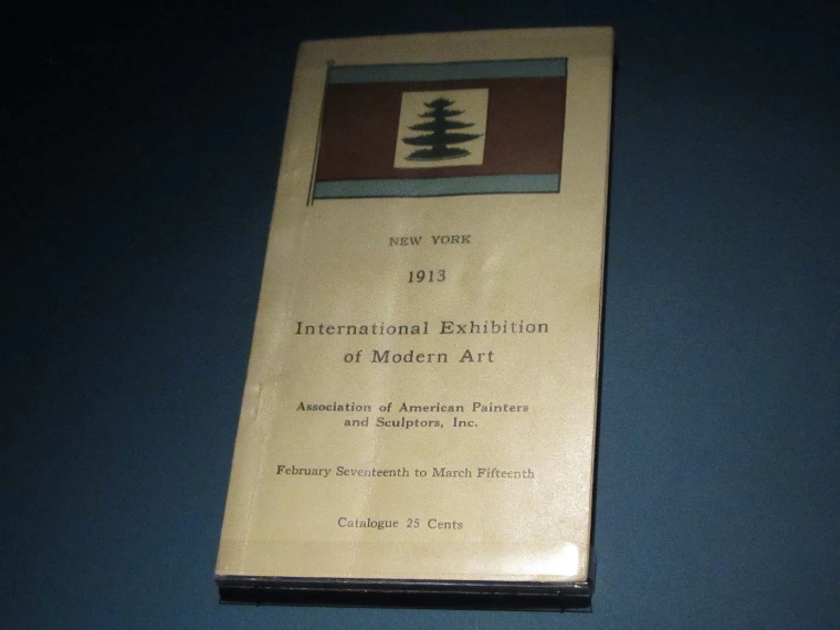 A cream paper catalogue for the 1913 International Exhibition of Modern Art, with a pine-tree flag on its cover, displayed in a museum case (2014).