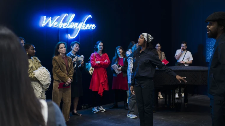 Visitors stand in a darkened space under a blue neon sign reading We belong here as a woman speaks beside a piano (2026).
