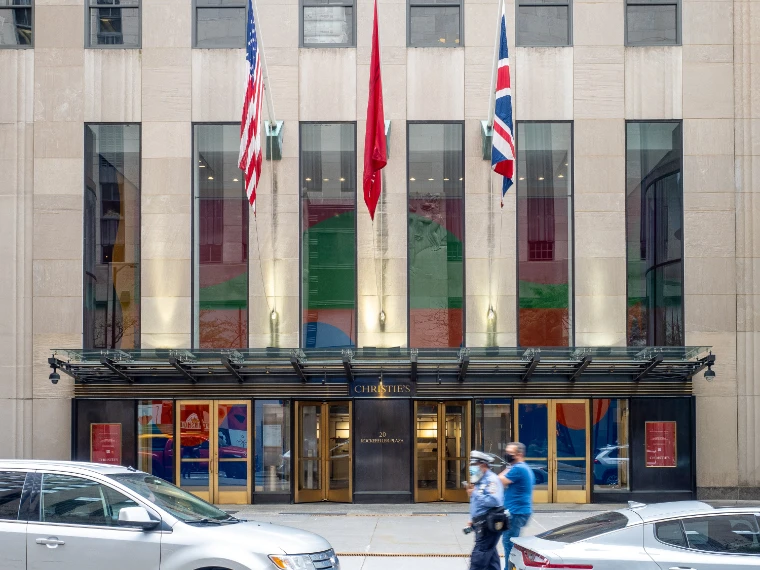 The limestone front of Christie’s at Rockefeller Center, flags flying over its canopied entrance and tall windows hung with coloured banners (2021).