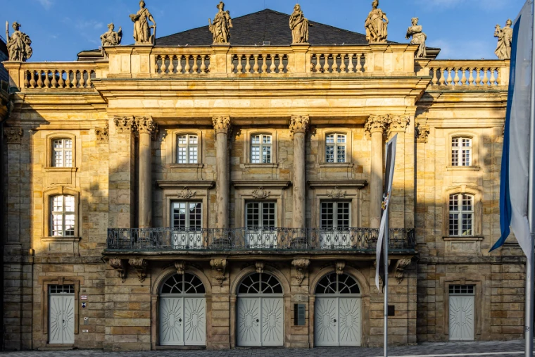 The sunlit sandstone facade of the Margravial Opera House, with columns, a wrought-iron balcony and statues along the roofline under a blue sky (2019).