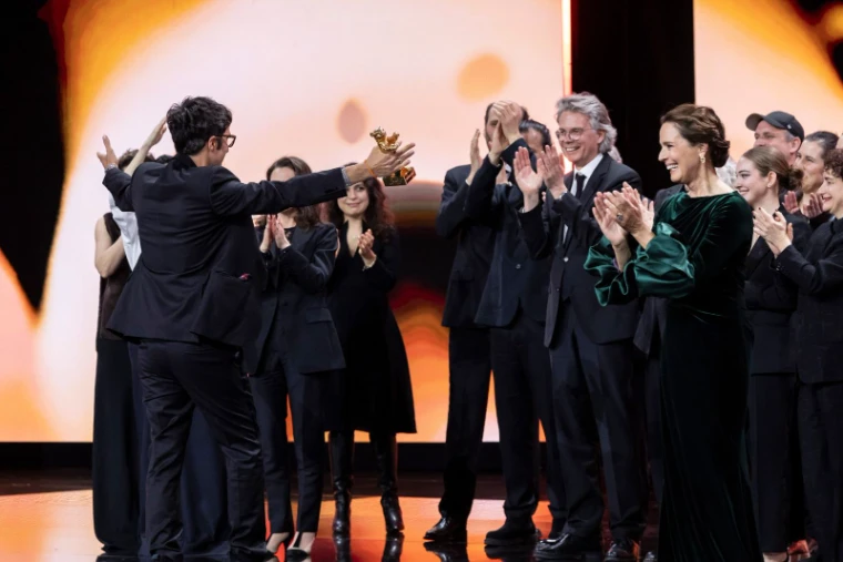 A director holding up the Golden Bear on stage as the jury and cast applaud at the award ceremony of The Berlinale, an engagement in the season calendar of The Mansa Court