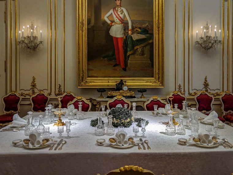 A long banquet table laid with white damask, crystal decanters and glasses, red-upholstered gilt chairs and a full-length imperial portrait on the wall beyond (2018).