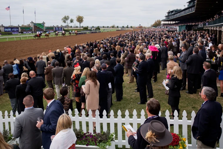 Racegoers in coats and hats crowded along a white picket fence watching the race on the dirt track, the grandstand behind.