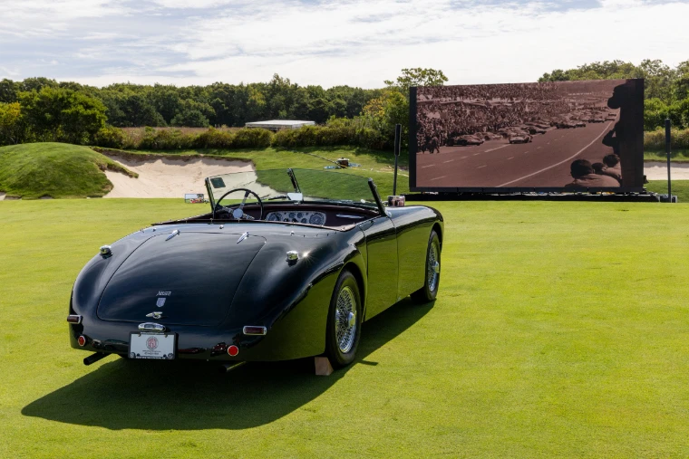 A dark green Allard roadster parked on the fairway faces a large outdoor screen showing black-and-white footage of a motor race start (2026).