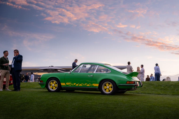 A green Porsche 911 Carrera RS with yellow script stands on the fairway at dusk under a pink and violet sky, guests talking around it (2026).
