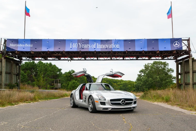 A silver Mercedes-Benz SLS with its gullwing doors open stands on the old track beneath a steel bridge hung with a “Mercedes-Benz 140 Years of Innovation” banner (2026).