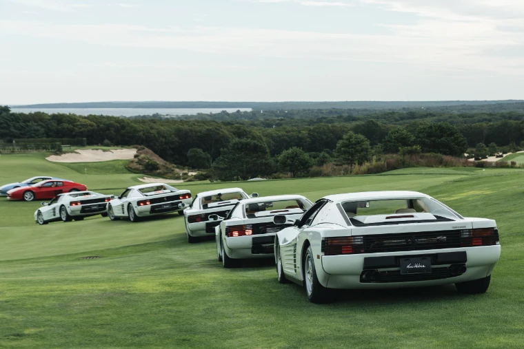 A row of white and red Ferrari Testarossas lined up on the grass of the driving range, with the bay beyond (2026).