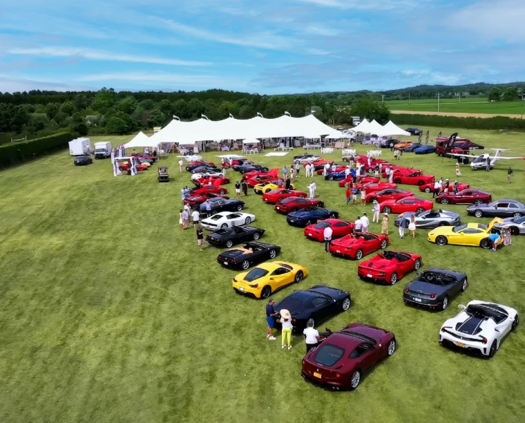 Rows of red, yellow and black sports cars drawn up on a lawn before a line of white marquees, seen from above, for The Concours Luncheon, Bridgehampton, New York, early June in the season calendar of The Mansa Court