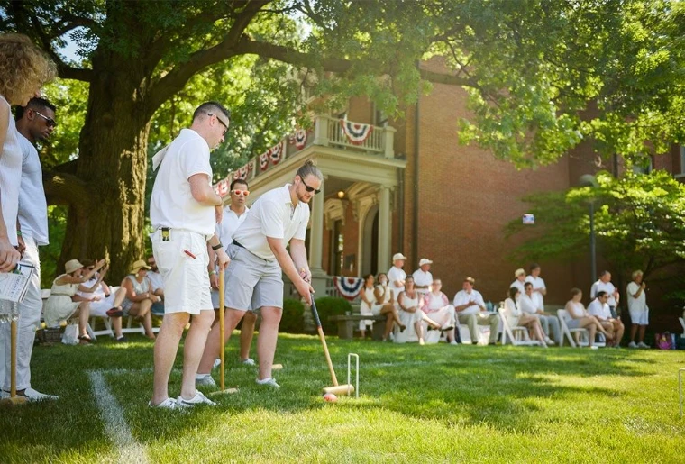 Players in whites at croquet on a lawn shaded by old trees, guests seated in the shade before a brick house hung with bunting, for The Croquet and Lawn Party, New York, an afternoon in the season calendar of The Mansa Court