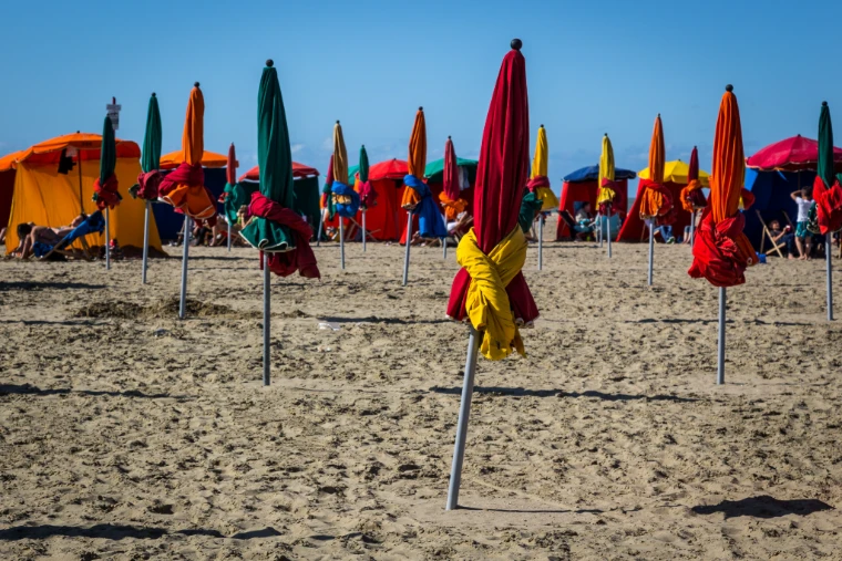 Furled parasols in red, yellow, orange and green stand in rows on the sand before bright beach tents under a blue sky (2014). Deauville beach, Normandy, France, shown for The Deauville August Meeting in the season calendar of The Mansa Court