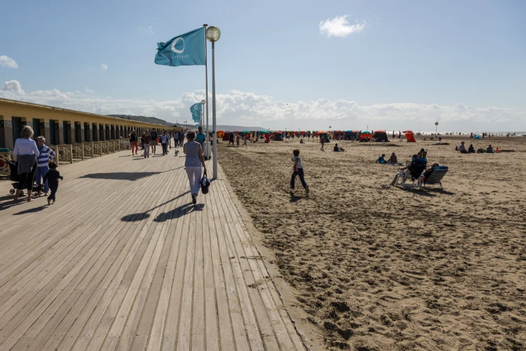 Strollers walk the wooden boardwalk beside the beach cabins and the sand under flags and a pale summer sky (2014). Les Planches, Deauville, Normandy, France, shown for The Deauville August Meeting in the season calendar of The Mansa Court