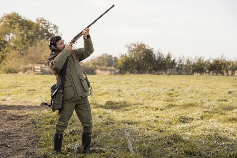 A man in tweeds and ear defenders raises a side-by-side shotgun beside a wooden peg numbered 3 in an autumn field (2018).