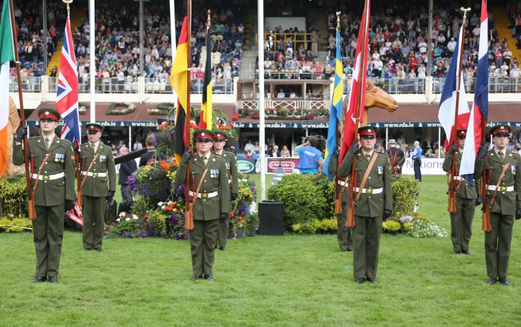 Soldiers in dress uniform stand in the arena before a row of national flags and a full grandstand (2012).