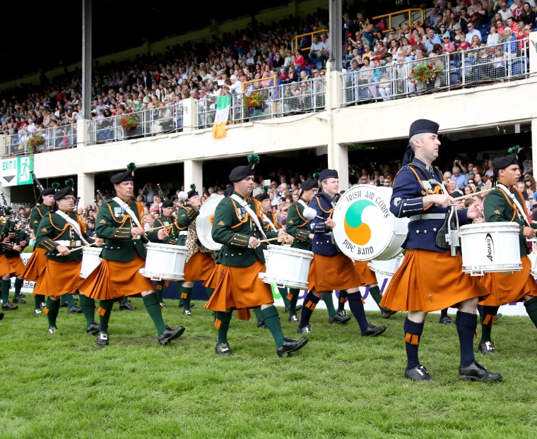 A military pipe band in saffron kilts marches across the grass in front of a packed grandstand (2012).
