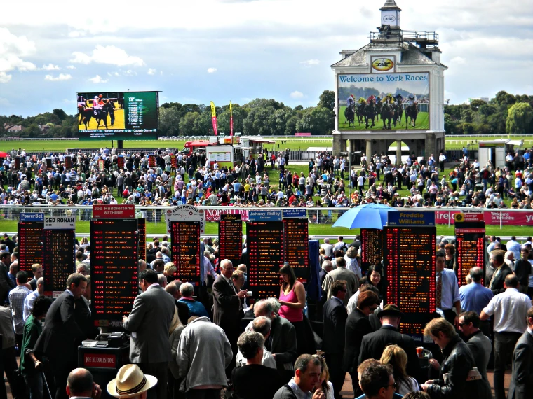 Racegoers crowd around bookmakers’ boards on the lawn, with the clock tower, big screen and track beyond under a summer sky (2010).